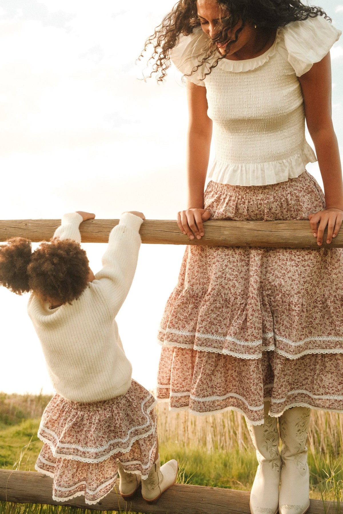 A woman and a young girl in matching white tops, boots, and floral skirts smile while climbing a wooden fence in a grassy field, showcasing the adorable Mini Annalise Skirt in Philocaly Floral by Ivy City Co for girls.