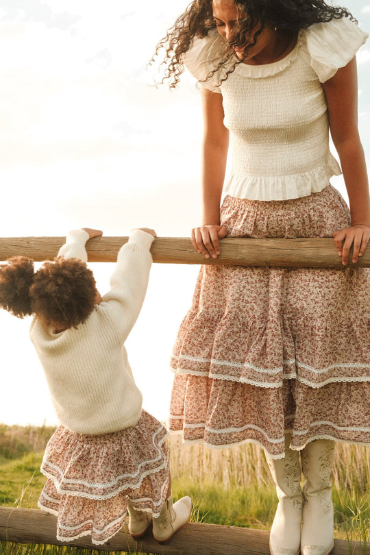 A woman and a young girl in matching white tops, boots, and floral skirts smile while climbing a wooden fence in a grassy field, showcasing the adorable Mini Annalise Skirt in Philocaly Floral by Ivy City Co for girls.