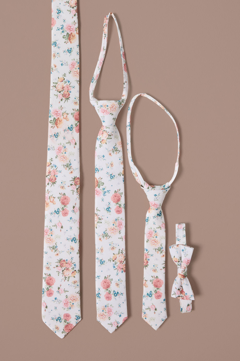 Four “Ties in Countryside Peony Blossom” by Ivy City Co rest on a brown background: two classic neckties, one skinny tie, and one bow tie—all featuring a white base adorned with multicolored floral patterns inspired by countryside peony blossoms—perfect for coordinating a charming family look.