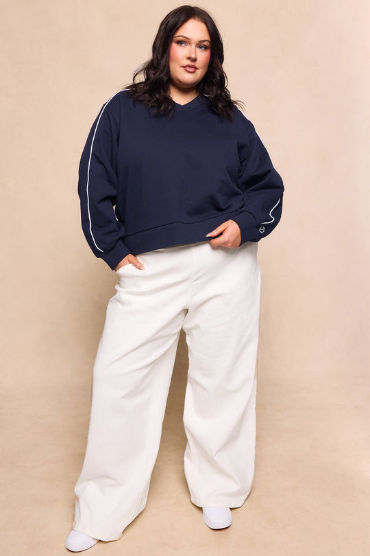 A woman poses confidently against a beige background wearing the Ivy City Co Kinsley Sweatshirt in navy, featuring white piping on the sleeves, styled with wide-leg white pants and white shoes.