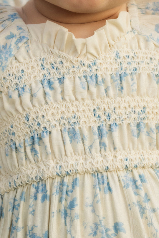 A close-up of a child wearing the Ivy City Co Baby Roselyn Romper in Ivory Porcelain Fleur, featuring smocked detailing and a ruffled neckline.