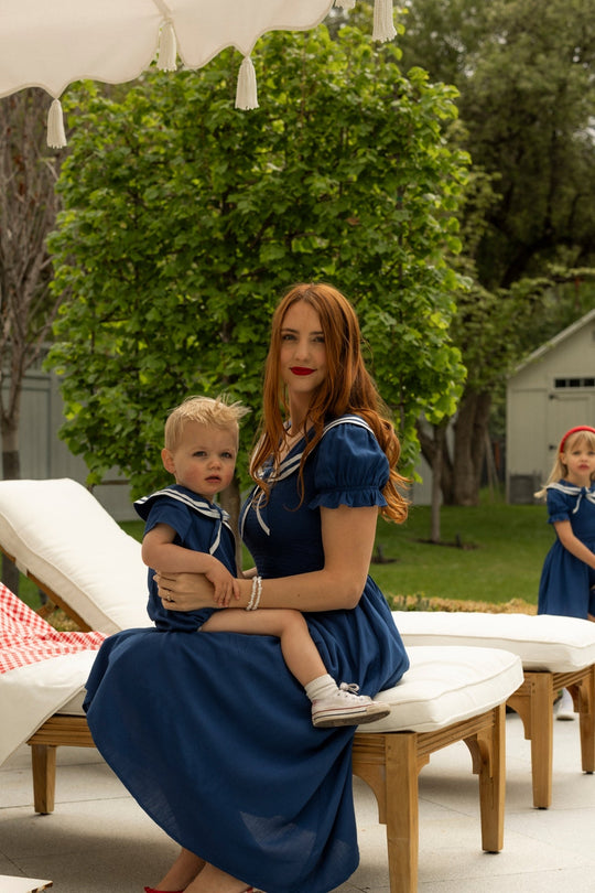 A woman in a long blue dress sits on a lounge chair outdoors, holding a young child dressed in the Ivy City Co Baby Sailor Romper in Navy. Another child, also wearing blue, stands in the background amidst the lush garden’s vibrant greenery.