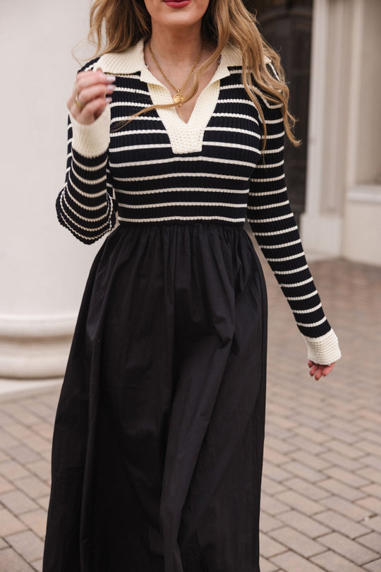 A woman is wearing the Ivy City Co Adelina Dress in Black, a black and white striped knit long sleeve dress with a collar, while holding a necklace. The photo is taken outdoors on a paved walkway, with her face partially out of frame.