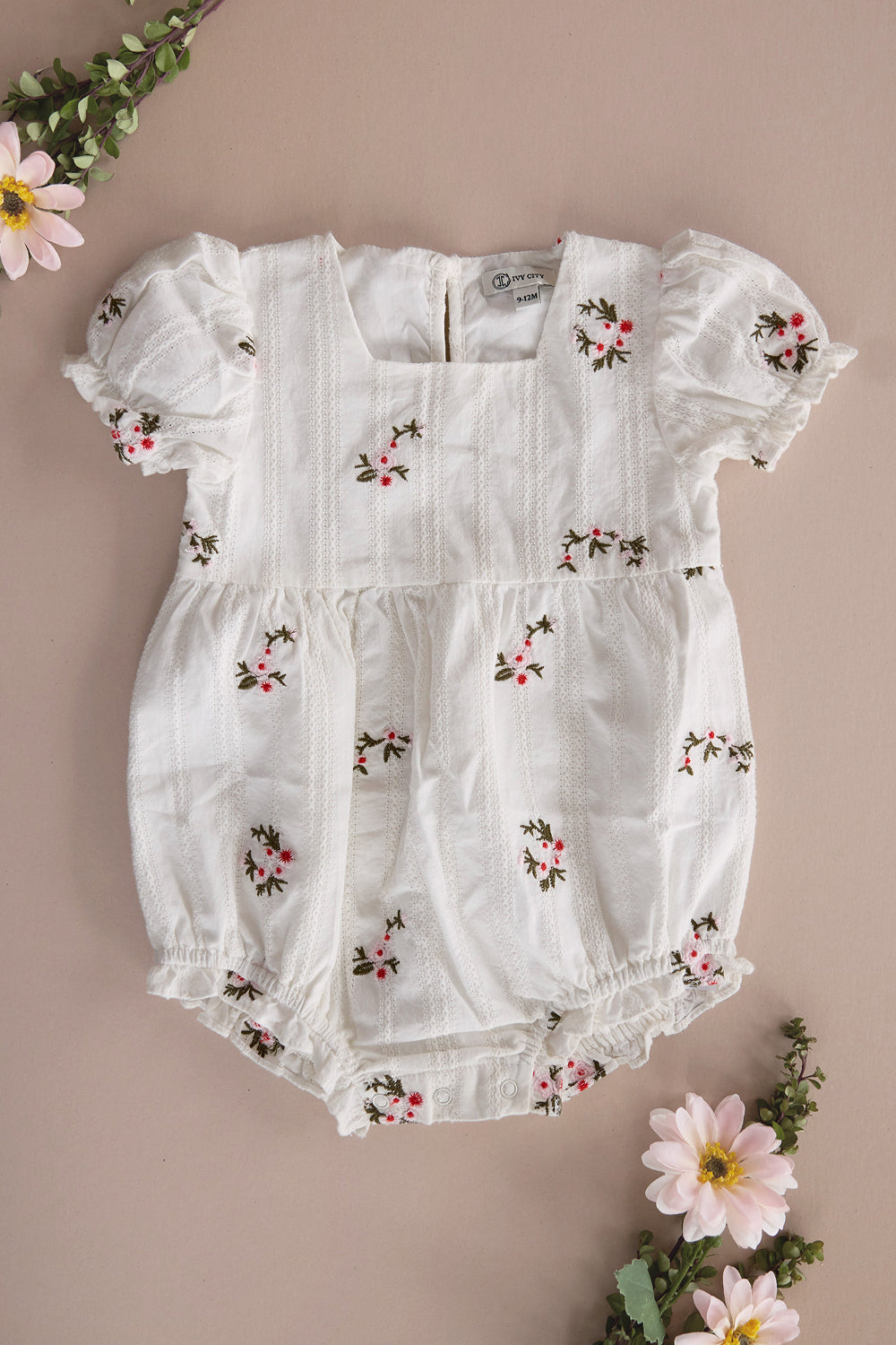 The Baby Jessie Romper in Ditsy Embroidery by Ivy City Co, featuring a smocked bodice, puff sleeves, delicate floral embroidery, and elastic leg openings, is displayed on a beige surface adorned with pink and white flowers and green foliage around the edges.