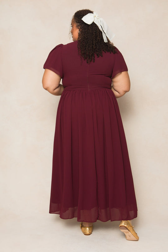A woman stands facing away, wearing the Abbie Dress in Wine Chiffon by Ivy City Co, featuring short sleeves and paired with gold shoes. Her hair is tied back with a large white ribbon against a soft beige background.