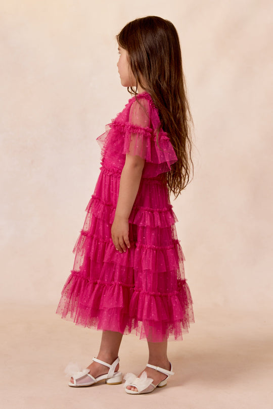 A young girl stands sideways, looking to the left against a light, neutral background, wearing the Ivy City Co Mini Whimsical Dress in Fuchsia with flutter sleeves and a layered tulle skirt, paired with white sandals.