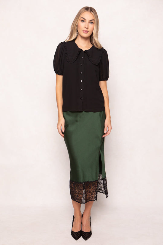 A woman with long blonde hair and a neutral expression stands against a plain background, wearing the Ivy City Co Andrea Blouse in black chiffon, styled with a green satin skirt featuring black lace trim and paired with black high heels.