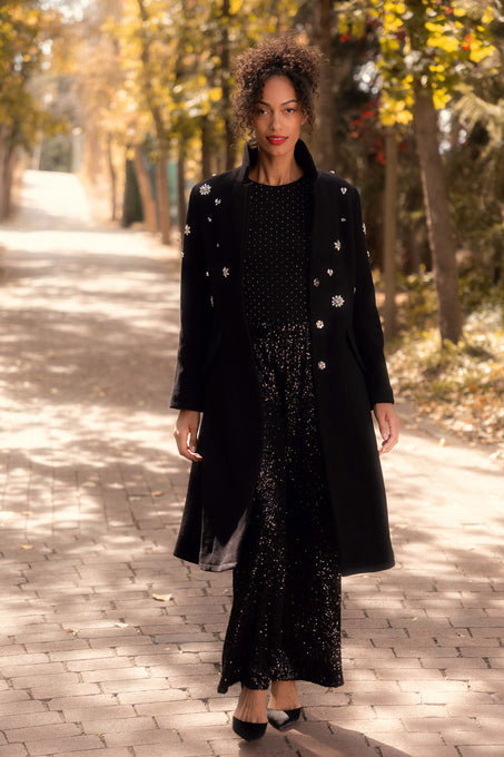 Woman wearing a black coat over a black dress with white polka dots, standing on a paved path with trees in the background.