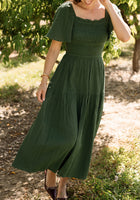 A woman strolling outdoors on a sunny day, surrounded by lush trees and greenery, wears the Jade Dress in Evergreen by Ivy City Co—a green, short-sleeve, smocked dress crafted from soft cotton. With one hand gently touching her hair, she showcases this stylish and maternity-friendly design that blends comfort with elegance for any occasion.