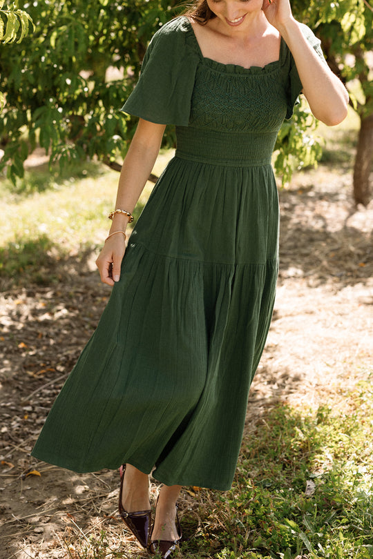 A woman strolling outdoors on a sunny day, surrounded by lush trees and greenery, wears the Jade Dress in Evergreen by Ivy City Co—a green, short-sleeve, smocked dress crafted from soft cotton. With one hand gently touching her hair, she showcases this stylish and maternity-friendly design that blends comfort with elegance for any occasion.