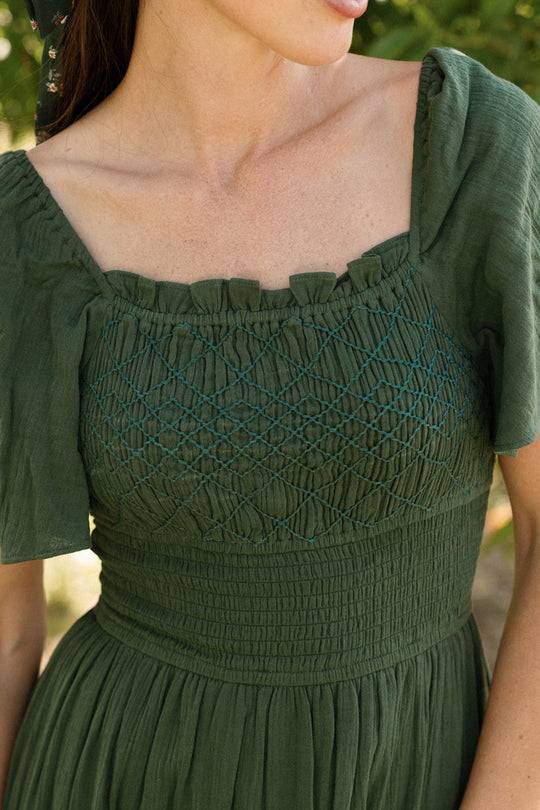 A woman stands outdoors wearing the Ivy City Co Jade Dress in Evergreen—a flattering, maternity-friendly cotton dress featuring puff sleeves and intricate smocked and embroidered detailing on the bodice.
