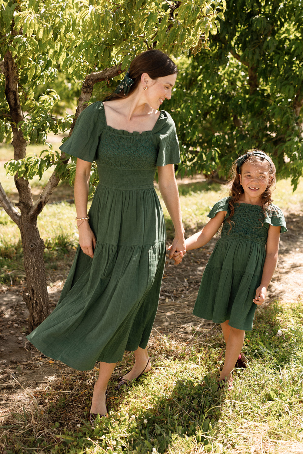 A woman and a young girl, both wearing matching Ivy City Co Jade Dresses in Evergreen, smile and hold hands while walking beneath sun-dappled trees on a grassy lawn. Their maternity-friendly outfits provide comfort and style, adding to the joy of the moment.