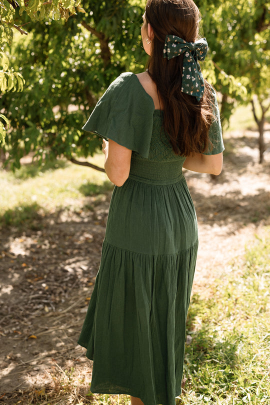 A woman is seen from behind in a sunlit orchard, wearing the Ivy City Co Jade Dress in Evergreen—a green, tiered cotton midi dress featuring flutter sleeves and a smocked waist. The look, which is maternity friendly, is perfectly finished with a floral bow hair accessory.