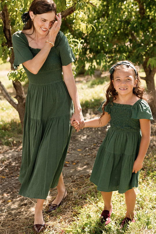 A woman and a young girl, both wearing matching Ivy City Co Jade Dress in Evergreen, hold hands and smile while walking outdoors in a sunny, leafy area. These maternity-friendly dresses from Ivy City Co make every moment together stylish and comfortable.