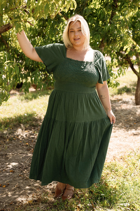 A woman wearing the Jade Dress in Evergreen by Ivy City Co stands outdoors beneath a tree, one hand gently holding a branch and the other grasping the edge of her maternity-friendly dress as sunlight filters through the leaves.