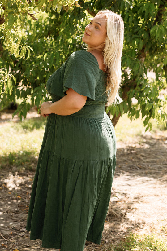 A woman with long blonde hair wears the Ivy City Co Jade Dress in Evergreen, standing outdoors amid lush green trees and sunlight, looking over her shoulder and smiling.