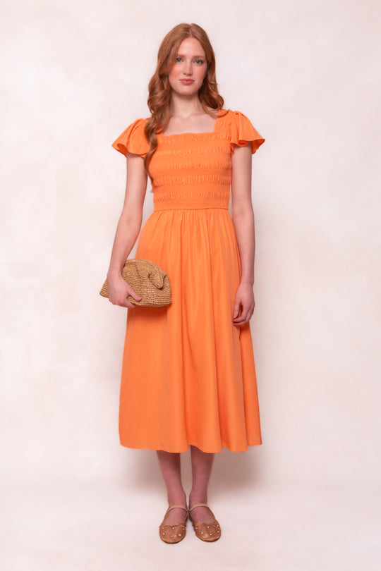 A woman with long, wavy red hair wears the Ivy City Co Janessa Dress in Orange, featuring a smocked bodice. She holds a woven clutch and wears tan flats, posed against a pale background.