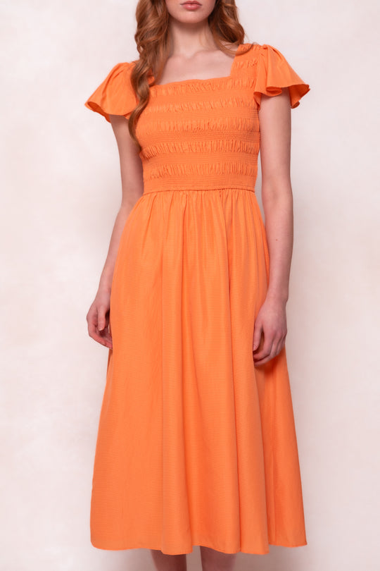 A woman wears the Janessa Dress in Orange by Ivy City Co, featuring seersucker fabric, a smocked bodice, short sleeves, square neckline, and gathered waist, standing against a light neutral background.