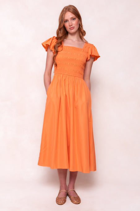 A woman with long red hair stands against a light background, hands in her pockets, wearing the Ivy City Co Janessa Dress in Orange—featuring ruffled sleeves and a smocked bodice—and brown flats.