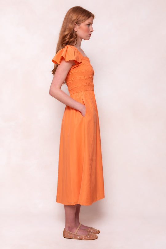 A woman with long red hair wears the Ivy City Co Janessa Dress in Orange, featuring a smocked bodice, short sleeves, and pockets. Posed in profile against a light backdrop, she pairs the dress with tan flats for a casual look.