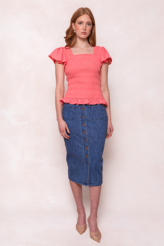 A woman with straight, light brown hair poses against a light background wearing the Ivy City Co Janessa Top in Pink—a ruffle peplum style with short sleeves, smocked details, and button front—paired with a knee-length denim skirt and pointed-toe heels.