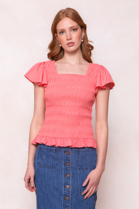 A woman with long, wavy red hair wears the Ivy City Co Janessa Top in Pink—featuring short sleeves, a square neckline, and a ruffle peplum—paired with a high-waisted blue button-front denim skirt, standing before a light background.