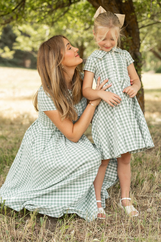 A woman and a young girl smile together outdoors under a tree, both wearing the Ivy City Co Amber Dress in Sage Gingham. The coordinating cotton midi dresses feature charming pleated skirts, adding a playful touch to their matching look as the woman kneels beside the girl.