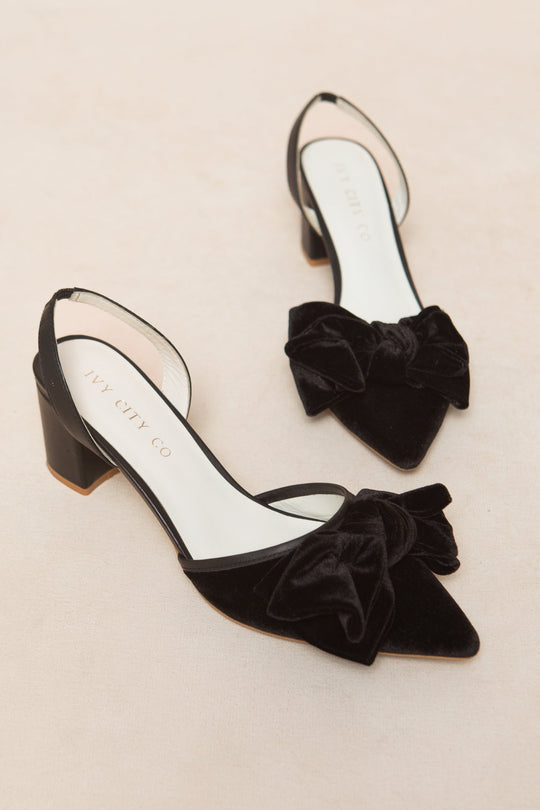 The Eloise Bow Heel in Black by Ivy City Co features a pair of black velvet, pointed-toe slingback shoes adorned with large black bows and sturdy block heels, displayed on a light beige surface.
