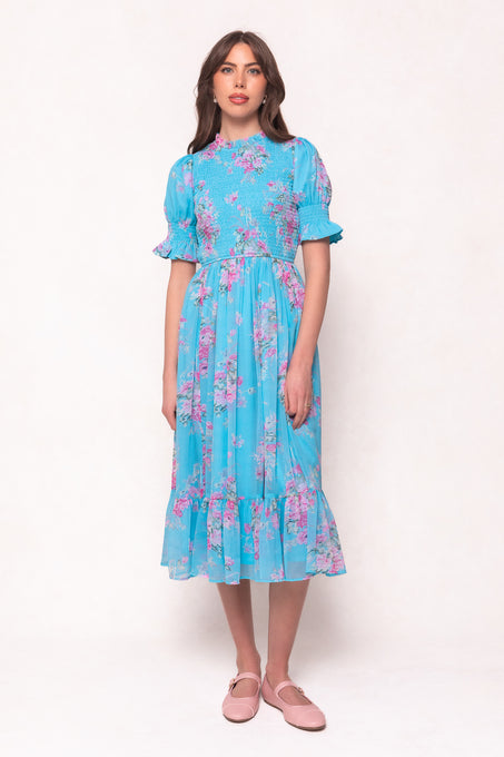 A woman wears the Ivy City Co Jennie Dress in Layla Floral—a bright blue dress with short puff sleeves and a smocked bodice—paired with light pink flats, against a plain background. This versatile style is maternity friendly.