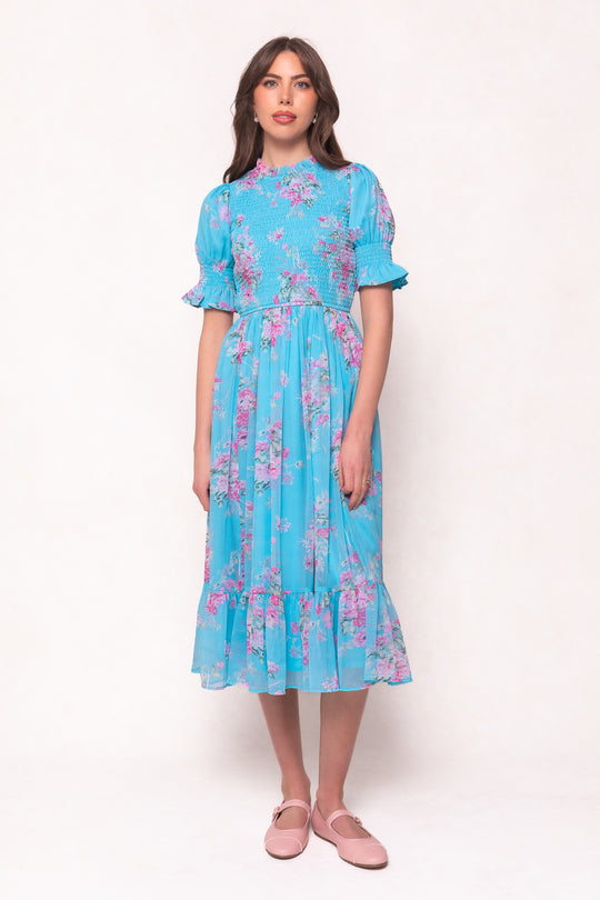 A woman wears the Ivy City Co Jennie Dress in Layla Floral—a bright blue dress with short puff sleeves and a smocked bodice—paired with light pink flats, against a plain background. This versatile style is maternity friendly.