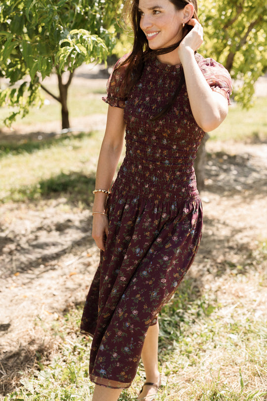 A smiling woman stands outdoors among trees on a sunny day, wearing the Ivy City Co Jennifer Dress in Aubergine Elysian Vine—a burgundy floral midi dress featuring puff sleeves and a flattering, maternity-friendly smocked bodice.