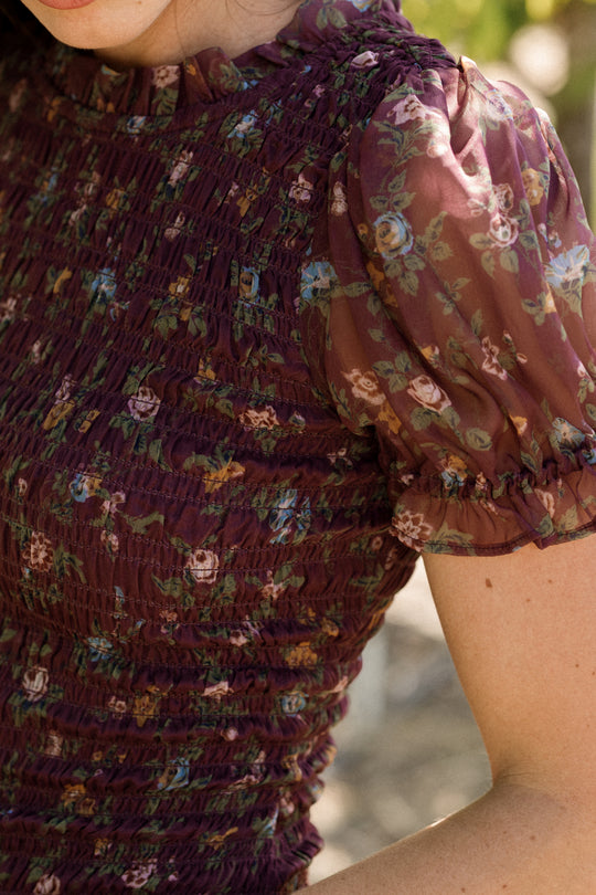 Wearing the Jennifer Dress in Aubergine Elysian Vine by Ivy City Co—a sheer, purple floral puff-sleeve dress with a smocked bodice that’s both stylish and maternity friendly—a person stands outdoors as sunlight highlights part of their arm and dress.
