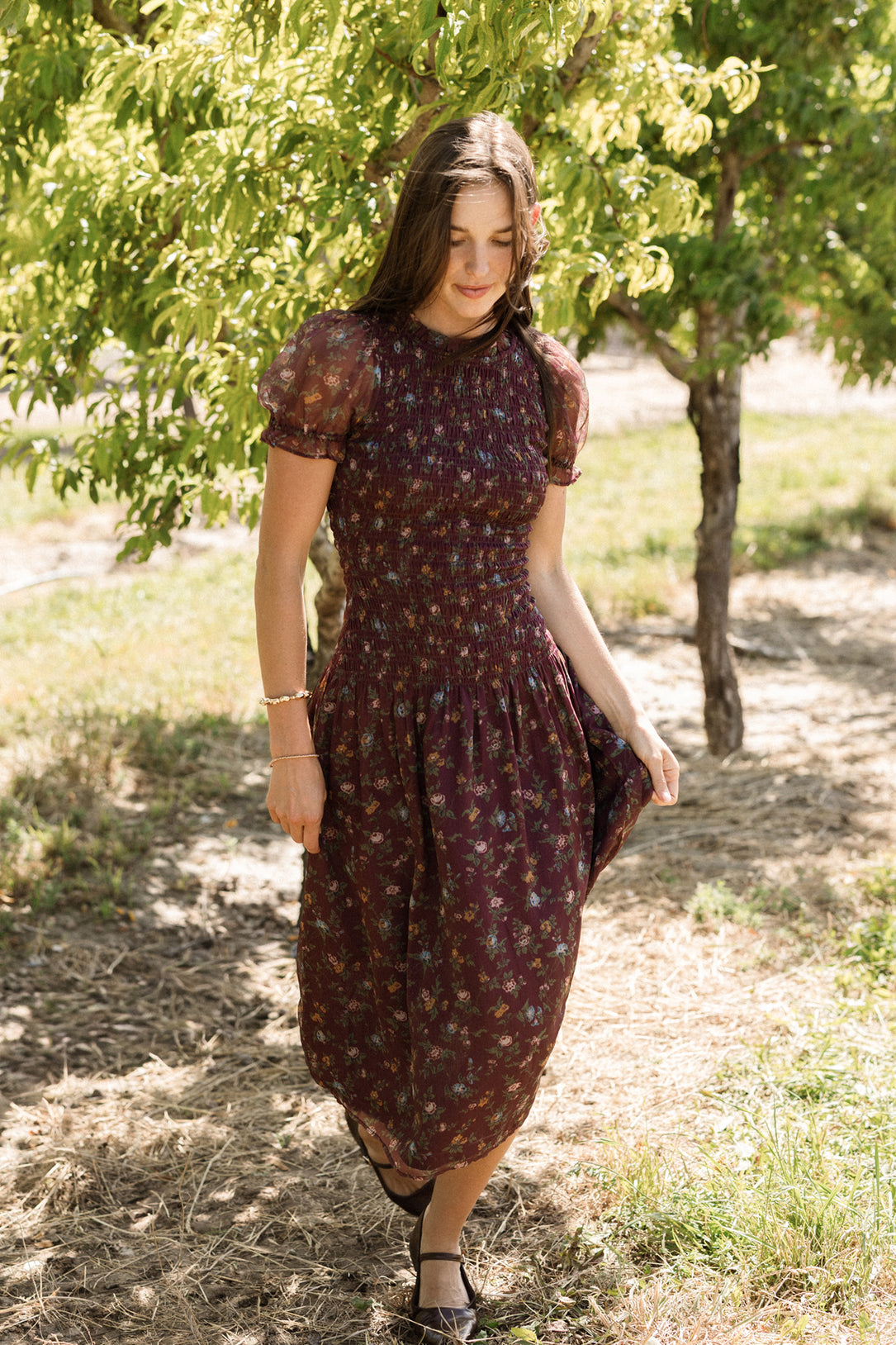 A woman in the Ivy City Co Jennifer Dress in Aubergine Elysian Vine, featuring a smocked bodice and dark floral print, walks outdoors on a sunny day, gracefully holding her skirt. Surrounded by lush green trees and gentle sunlight, she creates a serene, natural scene—showcasing an effortlessly elegant style that’s also maternity friendly.