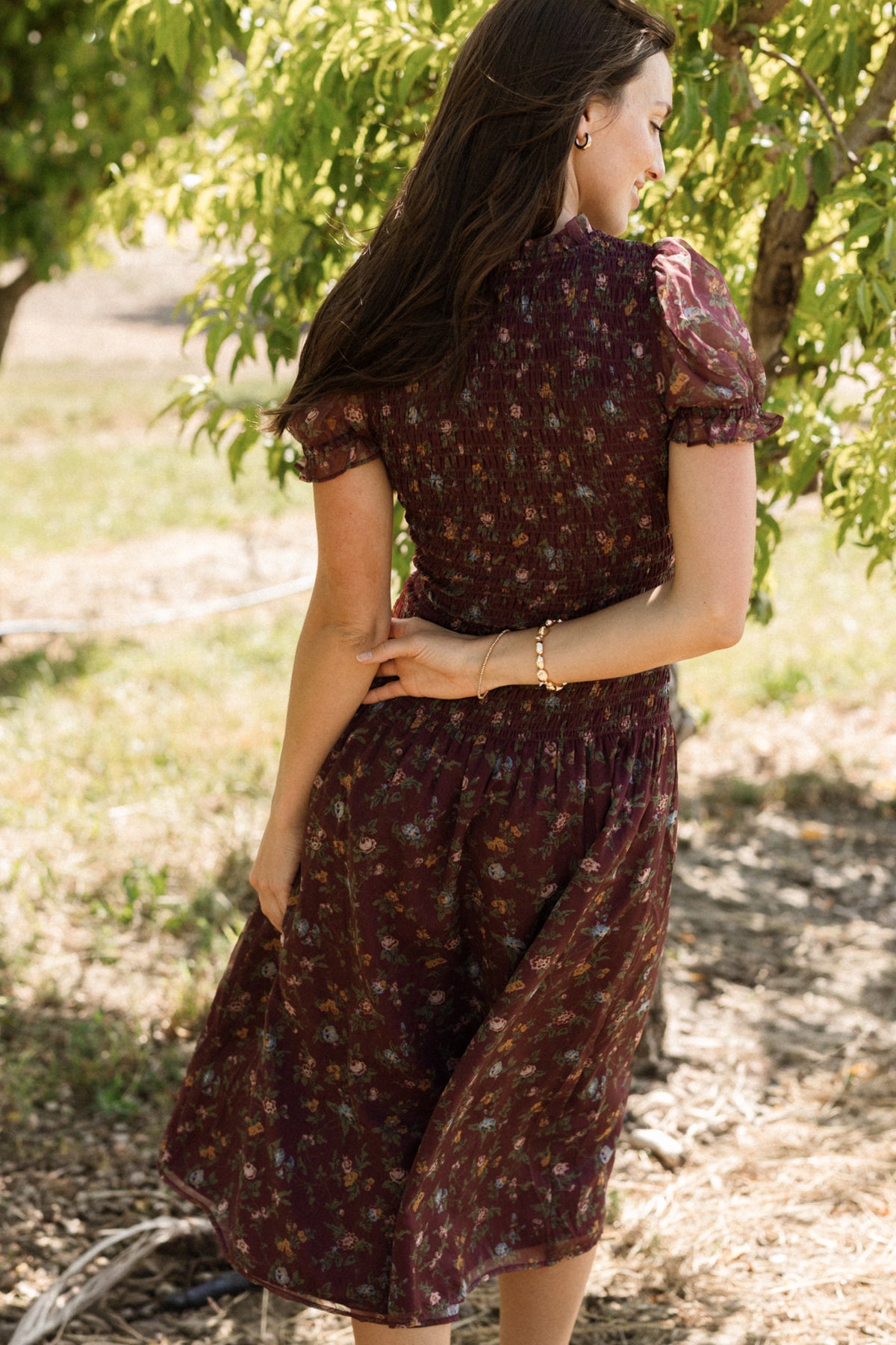 A woman in the Ivy City Co Jennifer Dress in Aubergine Elysian Vine, featuring a smocked bodice and floral design, stands outdoors surrounded by green trees as sunlight filters through the leaves. This dress is both stylish and maternity-friendly, making it perfect for enjoying comfort and elegance in nature.