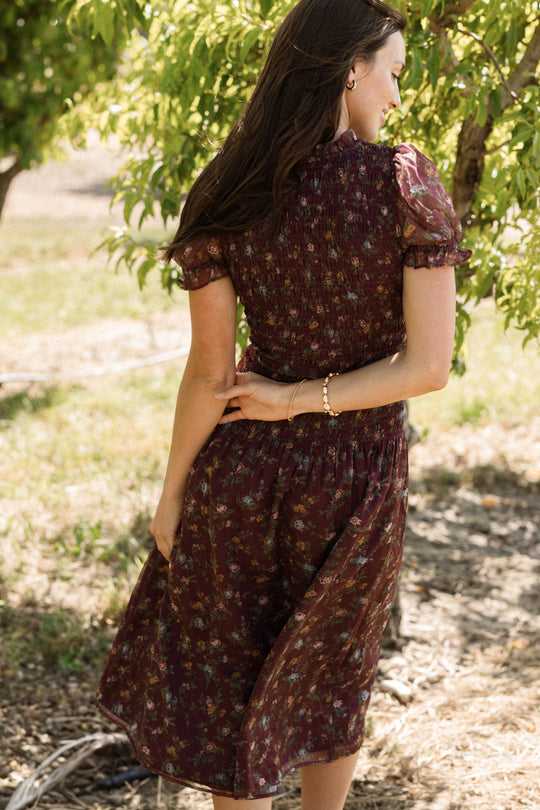 A woman in the Ivy City Co Jennifer Dress in Aubergine Elysian Vine, featuring a smocked bodice and floral design, stands outdoors surrounded by green trees as sunlight filters through the leaves. This dress is both stylish and maternity-friendly, making it perfect for enjoying comfort and elegance in nature.