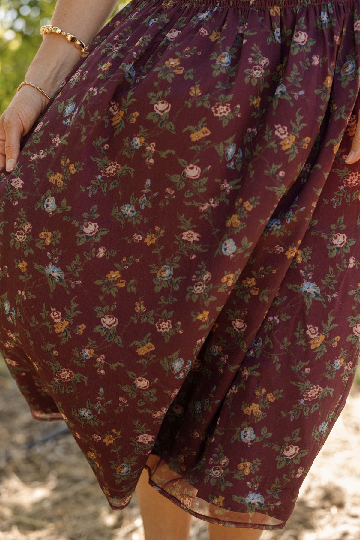 A person stands outdoors, their hand adorned with a gold bracelet and the skirt of their outfit visible—a piece reminiscent of the Ivy City Co Jennifer Dress in Aubergine Elysian Vine. The maroon fabric is decorated with small, colorful flowers, and is designed to be maternity friendly.