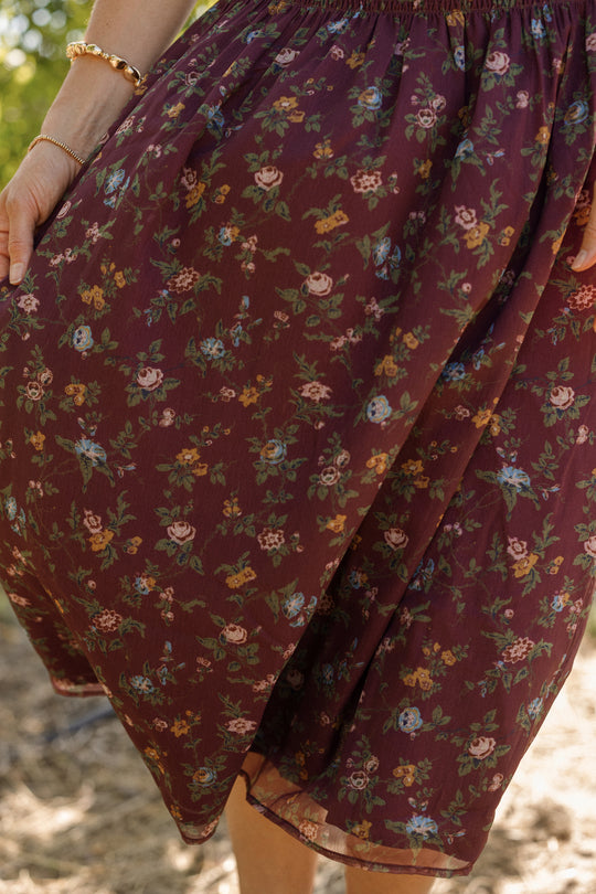 A person stands outdoors, their hand adorned with a gold bracelet and the skirt of their outfit visible—a piece reminiscent of the Ivy City Co Jennifer Dress in Aubergine Elysian Vine. The maroon fabric is decorated with small, colorful flowers, and is designed to be maternity friendly.