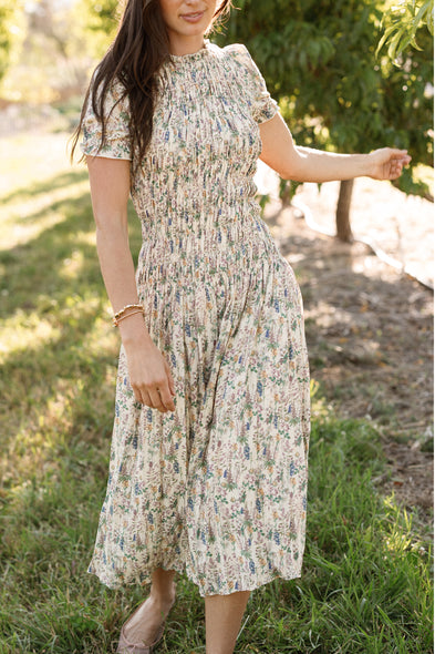 A woman stands outdoors on grass, barefoot and surrounded by trees and sunlight, wearing the Ivy City Co Jennifer Dress in Autumn Bouquet—a light-colored, floral-print, midi-length dress with a smocked bodice and gathered waist. Featuring short sleeves, a relaxed fit, and a maternity-friendly design, this style perfectly complements her long brown hair for an effortlessly graceful look.