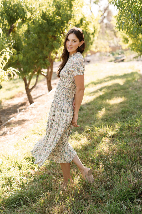 A woman wearing the Ivy City Co Jennifer Dress in Autumn Bouquet, featuring a smocked bodice and light floral pattern, stands sideways on the grass, smiling with one foot slightly lifted. Sunlight filters through leafy trees behind her, creating a soft, natural backdrop that highlights this maternity-friendly style.