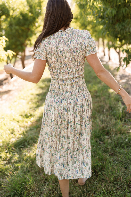 A woman wearing the Jennifer Dress in Autumn Bouquet by Ivy City Co, featuring a smocked bodice and a floral pattern, walks through a sunlit grassy orchard with her arms slightly outstretched, facing away from the camera. This maternity-friendly dress flows beautifully as she moves through the dappled light.
