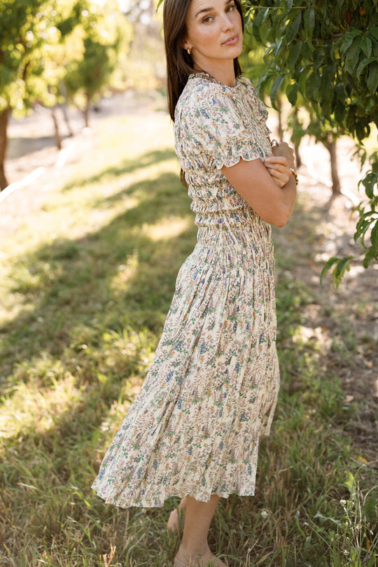 A woman stands outdoors in the grass next to a leafy tree on a sunny day, wearing the light-colored, floral-patterned Jennifer Dress in Autumn Bouquet by Ivy City Co. Featuring a smocked bodice and an elegantly maternity-friendly design, she looks at the camera with her arms crossed.