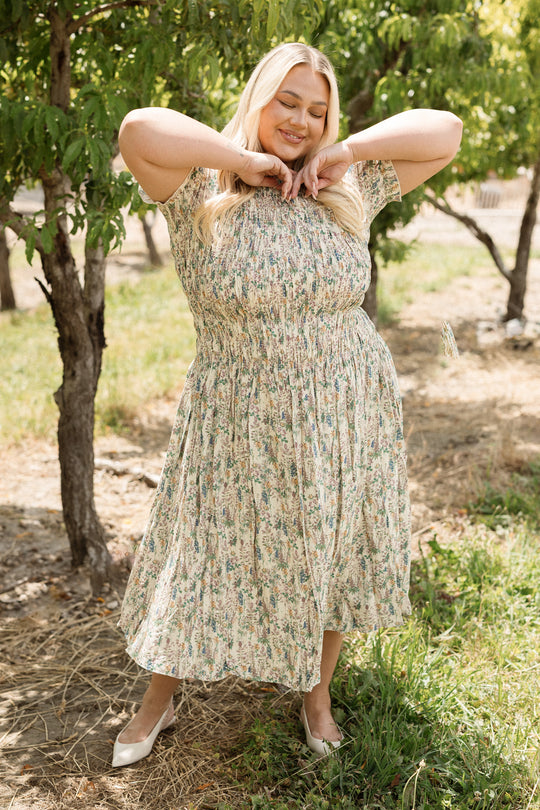 A smiling woman poses outdoors in a sunlit orchard, wearing the Jennifer Dress in Autumn Bouquet by Ivy City Co. With its smocked bodice and maternity-friendly design, this stylish dress is perfect for enjoying warm days surrounded by green trees and grass. She stands with her hands under her chin and eyes closed, radiating joy among nature.