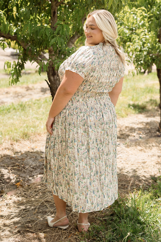 Wearing the Jennifer Dress in Autumn Bouquet by Ivy City Co, a woman stands on a sun-dappled dirt path surrounded by lush green trees, smiling as she looks over her shoulder. With its smocked bodice and floral print, this effortlessly maternity-friendly dress offers comfort and style for enjoying nature on a bright day.