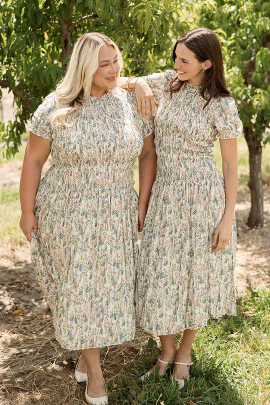 Two women stand outdoors under trees, smiling at each other. Both are wearing matching Ivy City Co Jennifer Dresses in Autumn Bouquet with a smocked bodice and white shoes. The sunlight creates a warm, cheerful atmosphere in this maternity-friendly scene.