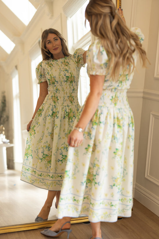 A woman admires her reflection in a bright room, wearing the Ivy City Co Jennifer Dress in Garden Tea Party Floral, featuring puff sleeves, a smocked waist, and breezy lightweight fabric.