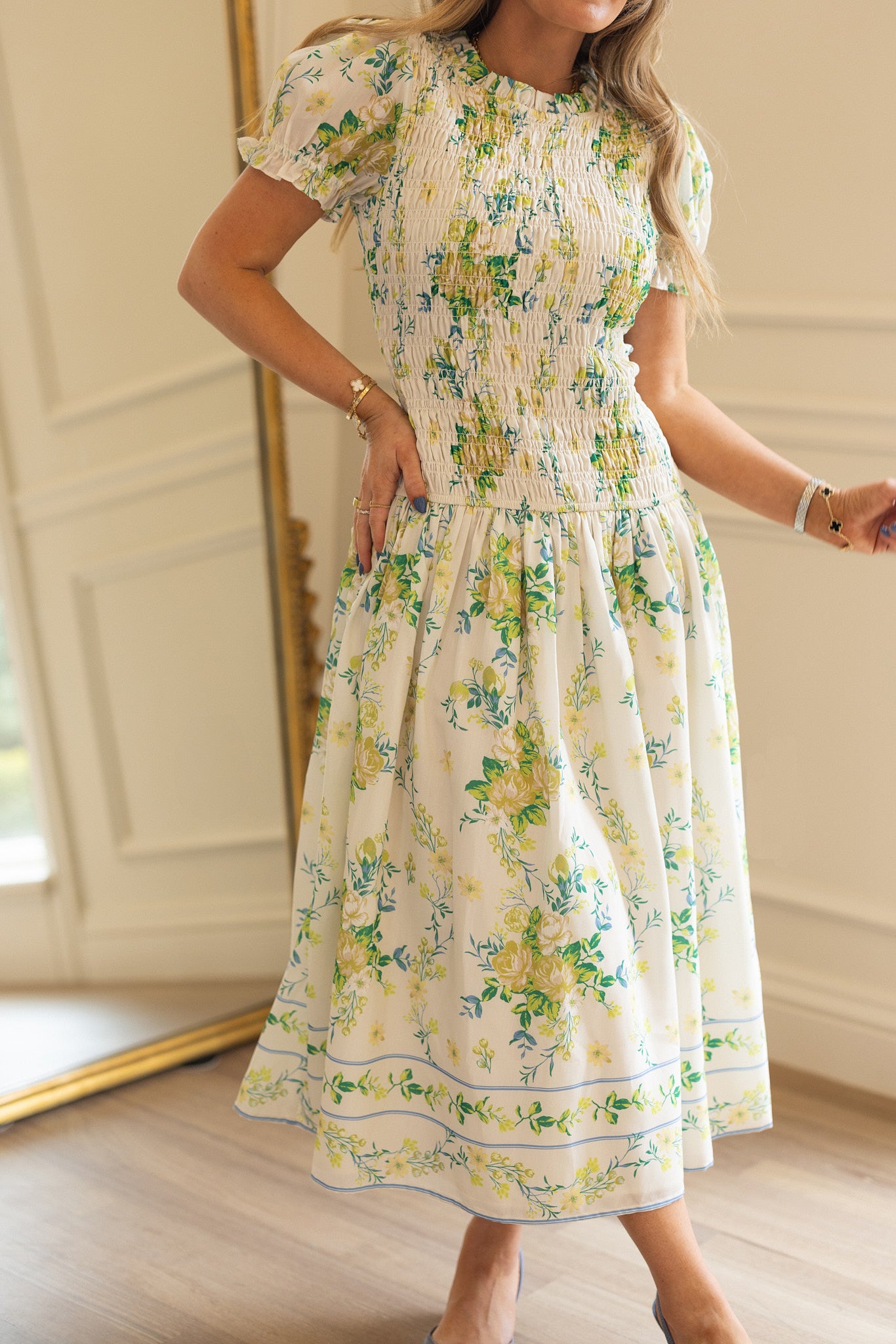 A woman wears the Ivy City Co Jennifer Dress in Garden Tea Party Floral, a white, short-sleeved, maternity-friendly dress with green and yellow blooms, standing indoors by a large gold-framed mirror.