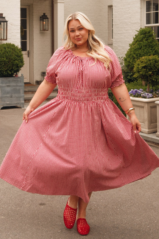 A woman with long blonde hair smiles outdoors, holding the sides of her Ivy City Co Jessie Dress in Red Gingham, with greenery and a white building in the background.