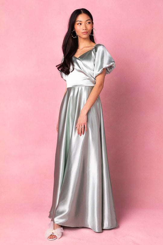 A woman stands against a pink background, wearing the Ivy City Co Tessie Dress in Sage—a shiny silver, floor-length gown featuring short puff sleeves—and white open-toe shoes, gazing slightly to the side. This modest bridesmaid dress exudes elegance and timeless sophistication.