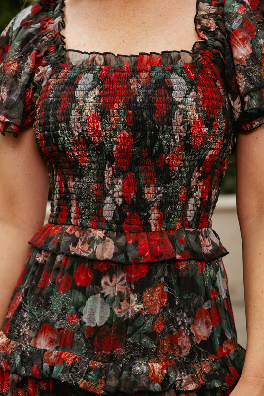 A person is shown wearing the Ivy City Co Winnie Dress in Midnight Roses, featuring a square neckline, puff sleeves, and ruffled layers with a red and green floral pattern. The smocked, textured fabric creates a beautifully maternity-friendly fit. Only the upper torso and part of the arm are visible.