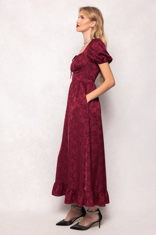A woman in a side profile pose wears the Amira Dress in Red by Ivy City Co, a jacquard maxi dress with puff sleeves, a ruffled hem, pockets, and a fitted bodice. She pairs the ankle-length dress with black ankle-strap heels. The background is plain and light.